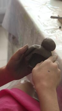 Close-up of a child shaping small cylindrical pieces of clay on a work surface, with tools and water marks visible. The scene captures the early stages of a clay modeling project, emphasizing