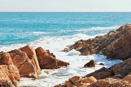 Rocky coastline with waves and blue sea. Waves crashing on brown rocky shore. Seascape with rugged rocks and ocean. Coastal scene of rocks and blue water. Rocky shoreline meeting turquoise sea