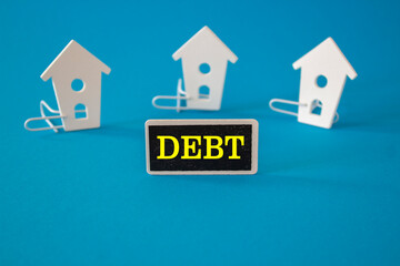 DEBT symbol. Wooden houses stay next to a wooden black board with the words DEBT. Beautiful blue background. Concepts of liabilities and taxes that must be paid.