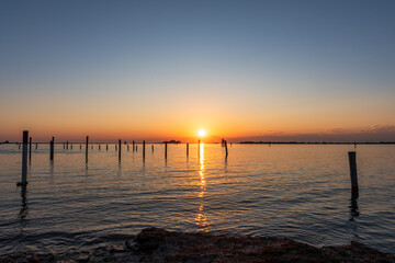 Golden sunset over the calm waters in Grado, Italy, with a line of wooden poles reflecting the last light of day in a peaceful summer scene.