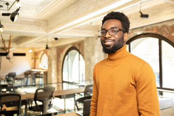 Confident young African man in glasses smiling and standing in modern office