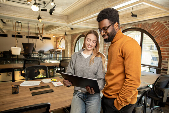 Diverse colleagues discussing project while standing in modern loft office and reviewing document