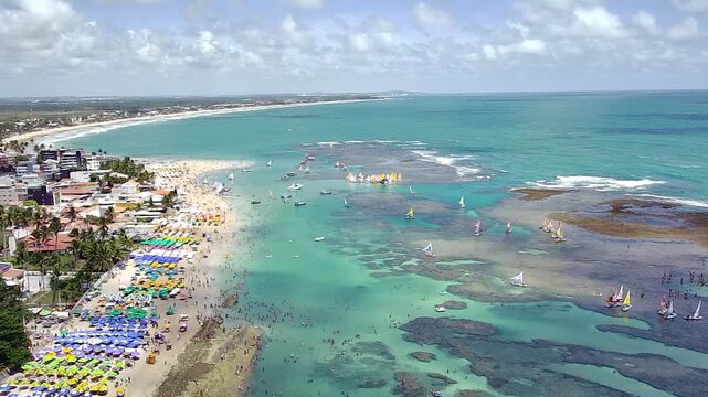 Aerial view of Pontal do Cupe Beach in Porto de Galinhas, Ipojuca, Pernambuco, Brazil. Sunny day with tourists, colorful umbrellas, sailboats, and natural pools in the turquoise sea.