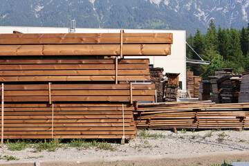 Stacked timber planks at a sawmill with forest and mountain in the background. Outdoor storage of sawn wooden planks at a lumber yard with scenic forest and mountains in the background