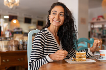 A happy moment in a cozy, warm cafe, where a woman is enjoying a delicious piece of cake with a fork. She smiles, savoring every bite