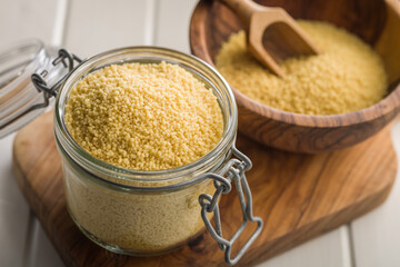 Raw couscous in jar on wooden cutting board.
