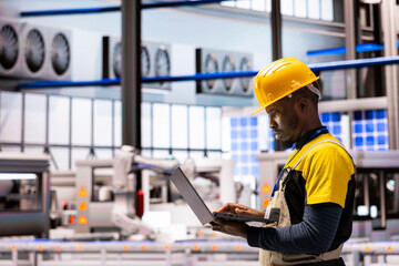 Engineer in smart manufacturing plant checks performance of automated systems on laptop. Integration of robotic arms and high tech machinery showcases Industry 4.0 advancements, optimizing production.