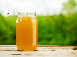 Glass jar of golden bee honey is on a wooden table, green grass field and sky in the background. Fine organic nature product concept. Agriculture and food industry.
