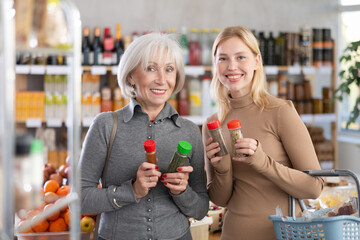 Girl with senior female woman pick up seasoning in store. Customer consider make choice between chopped dry culinary plants, compare varieties products. Client hesitate before buying, choose spices