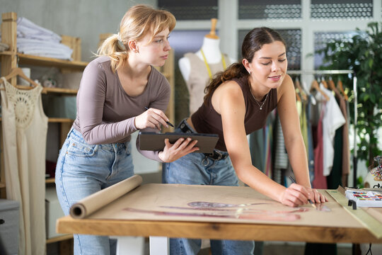 Female tailors work together to create patterns for clothes, they draw on paper and develop designs on a tablet. Fashion designers in the process of clothing production
