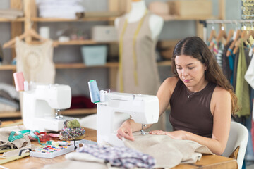 Female fashion designer sits at a table and works on a sewing machine, making straight stitches on...
