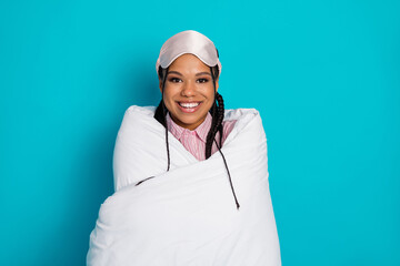 Cheerful girl wrapped in a warm duvet wearing sleepwear and a sleeping mask, smiling, on a blue background