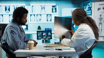 African american young man and specialist review ct scans and x rays, discussing about treatment after established diagnosis. Medic practitioner does scan interpretation, expertise. Camera B.