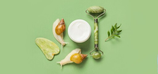 Snails, facial massage tools and cosmetic cream on green background, top view