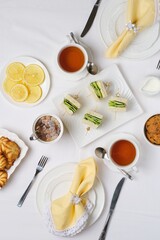 Tea setting for two with white tableware and yellow napkins, cucumber sandwiches and oatmeal cookies on a light tablecloth, top view.