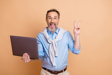 Elderly businessman holding laptop and showing peace gesture on beige background, stylish and cheerful expression