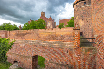 The Malbork Castle, a brick Gothic castle in northern Poland, Europe. Imposing red brick castle architecture with high walls, turrets, and towers under a dramatic, cloudy sky. Historic fortress.