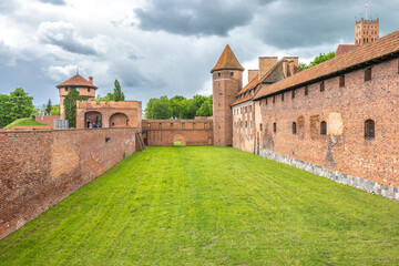 The Malbork Castle, a brick Gothic castle in northern Poland, Europe. Courtyard View of Historic Fortress: Brick walls, lush grass, towers under cloudy skies create an imposing architectural scene.