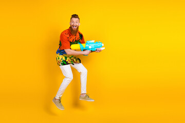 Cheerful young man in tropical shirt holding water gun on bright yellow background, conveying summer fun and holiday vibes