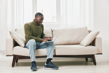 Young African American man smiling while working on a laptop at home, wearing a cozy sweater, showcasing a relaxed atmosphere in a bright living room