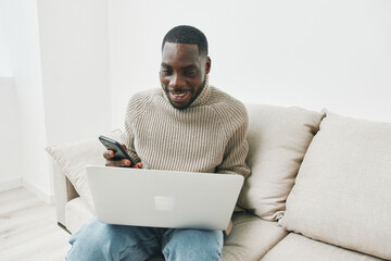 Young African man smiling while using smartphone and laptop on a comfortable couch in a bright living room, showcasing modern home lifestyle