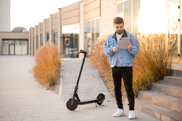 Young and handsome man standing with electric scooter outdoor and holding tablet device. Driving an ecological mode of transport.