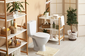 Interior of beautiful restroom with ceramic toilet bowl, houseplants and shelving unit near beige...