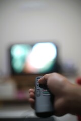 Hand holding TV remote, blurred screen in background. Leisure, entertainment, home relaxation, controlling media, technology use in modern living.