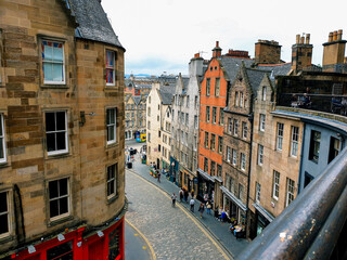 Obraz premium View of the Royal Mile in Old Town with a curved cobblestone street, colorful facades, and historic stone buildings under a cloudy summer sky.