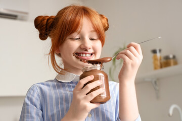 Cute girl with smeared face eating sweet chocolate paste from jar in kitchen
