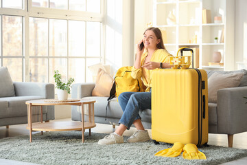Beautiful young woman with suitcase, headphones and backpack talking by phone in living room. Traveling concept