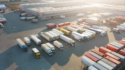 Aerial view of modern logistics terminal with semi-trailer trucks and cargo containers at sunset during golden hour at distribution center - Powered by Adobe