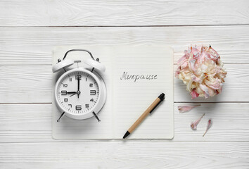 Notebook with word MENOPAUSE, flower and alarm clock on white wooden background