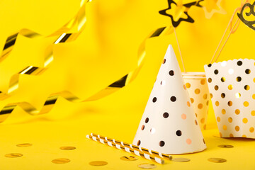 Composition with party hat, carton cups, box and confetti on yellow background