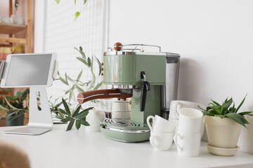 Modern coffee machine, cups, houseplants and tablet computer on table in kitchen
