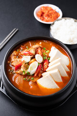 Korean food (Kimchi Jjigae), Kimchi soup with tofu, pork, scallion and chili in Korean stone bowl eating with cooked rice and kimchi cabbage on black background