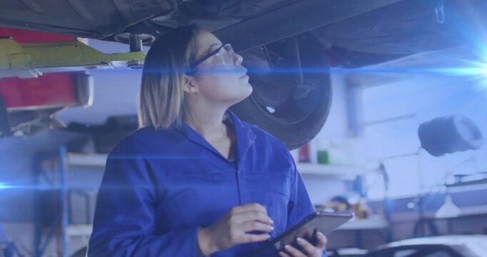 Female mechanic inspecting under lifted car in auto garage tapping tablet and logging diagnostics