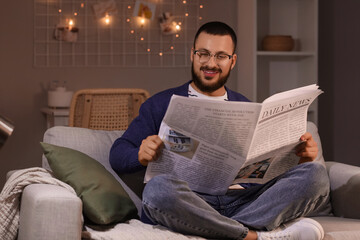 Fototapeta premium Young man with eyeglasses reading newspaper on sofa at home in evening
