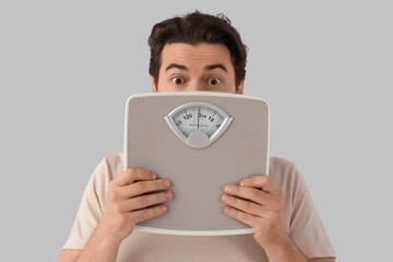 Shocked young man with weight scales on light background, closeup