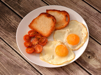 Two fried eggs, wheat toasts and fried cherry tomato halves on a wooden table.
