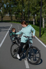 Fototapeta premium Caucasian woman riding bicycle with toddler son sitting behind her in child seat in park. Vertical photo. 