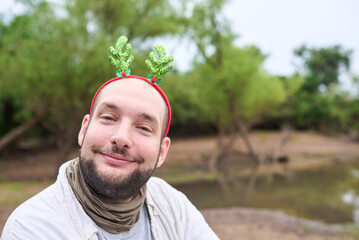 A young Hispanic man wearing a Christmas reindeer antler headband smiles at the camera outdoors, in a natural setting. © Carolina Jaramillo