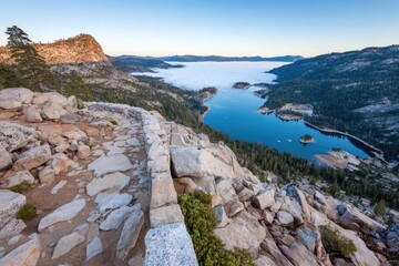 High-altitude vista of a serene lake nestled in a valley, framed by rugged mountains, with a stone path leading to the edge of the precipice