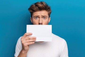 Attractive young man covering his mouth in shock over a solidarity message on paper against a blue background conveying fear and embarrassment