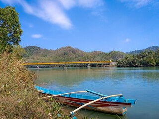 Fototapeta premium Traditional fishing boat on the riverbank with a yellow bridge and green hills in the background under a bright blue sky. Perfect for themes of nature travel, tropical rivers, Indonesian destinations