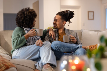 African American couple chatting on light grey sofa in living room holding ceramic mugs