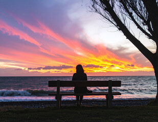 Silhouette of woman sitting on bench by the sea under tree watching vibrant sunset with golden light and serene coastal view