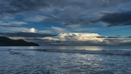 Obraz premium Early morning. Twilight. The ocean waves roll towards the shore, foam, and spread out. The clouds in the blue sky are illuminated with golden light. A hill in the distance. Malaysia. Borneo. 