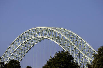 Steel arch of the Silver Jubilee Bridge rises above treetops, framed by a cloudless blue sky in bright sunlight