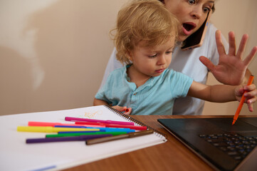 A mother manages remote work on her laptop while tending to her inquisitive toddler in a cozy home setting. The image captures multitasking and the challenges of work-life balance.